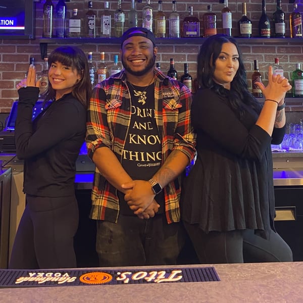 Three smiling O Grizzly's Bar & Bistro bartenders pose playfully behind a well-stocked bar.