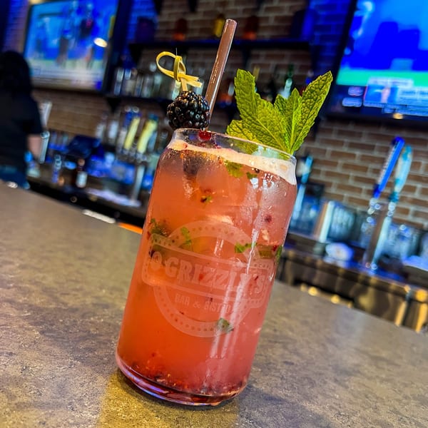 A red cocktail with mint and a blackberry garnish, branded with O Grizzly's logo, on a bar.