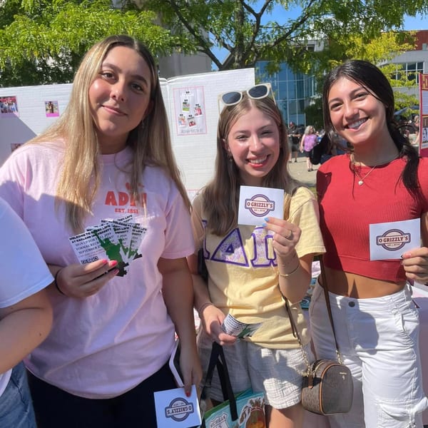 Three young women smiling and holding O Grizzly's Bar & Bistro flyers outdoors.