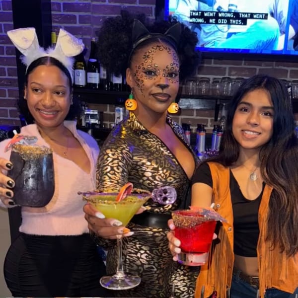 Three women in Halloween costumes holding colorful cocktails at O Grizzly's Bar & Bistro.